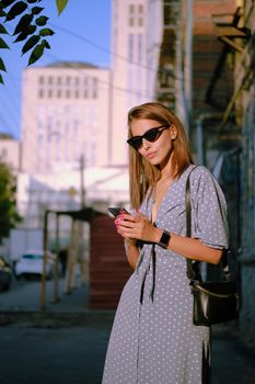 Wonderful blonde female in a long blue dress with polka-dots, watch, sunglasses, with a pendant around her neck and a small black handbag on her shoulder is using her smartphone while walking alone in the city. The concept of fashion and style. Close-up shot.