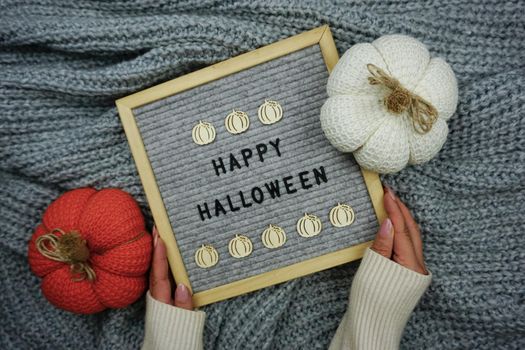 On a knitted gray background, a sign with the inscription - happy Halloween and there are knitted pumpkins. Women's hands hold a sign. Knitted pumpkins for Halloween celebration. Knitted decor.