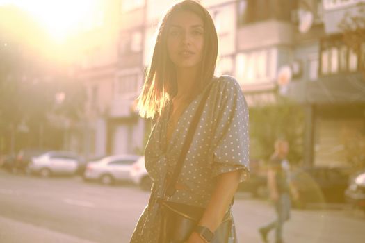 Beautiful blonde maiden in a long blue dress with polka-dots, watch, with a pendant around her neck and a small black handbag on her shoulder is looking at the camera while walking alone in the city. Sun flare. The concept of fashion and style. Close-up shot.