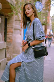 Soft Focus. Alluring blonde lady in a long blue dress with polka-dots, watch, with a pendant around her neck and a small black handbag on her shoulder is looking at the camera while posing standing sideways, walking alone in the city. The concept of fashion and style. Close-up shot.