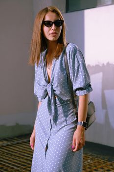 Beautiful blonde girl in a long blue dress with polka-dots, watch, sunglasses, with a pendant around her neck and a small black handbag on her shoulder is looking away while walking alone in the city. The concept of fashion and style. Close-up shot.