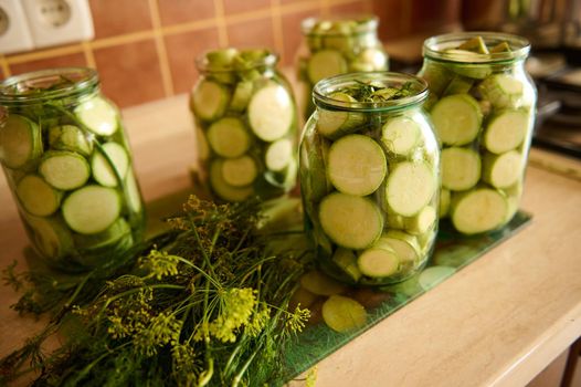 Sterilized glass jars with pickled slices of ripe organic vegetable marrow, marinated in salt or vinegar brine and fresh dill on kitchen table. Food preservation. Canning. Preserving for the winter