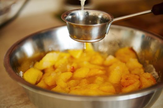 Close-up of pouring lemon juice into a metal bowl with sliced peaches while preparing homemade peach confiture. Canning peach. Preparing delicious jam. Making preserves for the winter. Selective focus