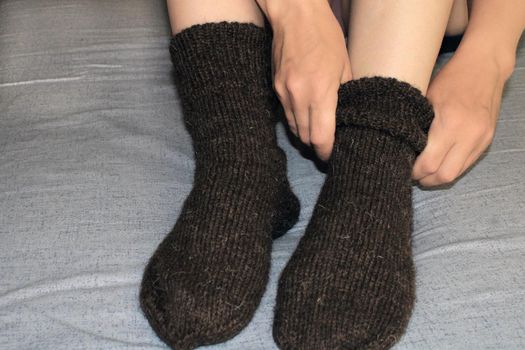 A young woman with slender legs sits on the bed at home and puts on woolen socks. The concept of the world energy crisis.