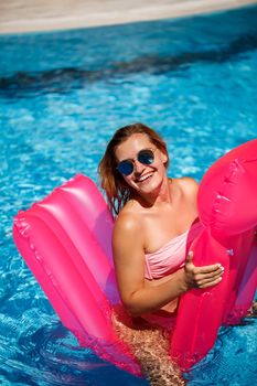 Sexy woman in sunglasses with a smile on her face in a swimsuit lies on a pink inflatable mattress in the pool. Relax by the pool on a hot summer sunny day. Vacation concept
