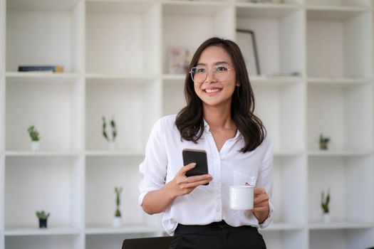 Asian businesswoman sitting at office holding coffee cup and smartphone