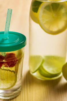 vertical photo of bottle and glass with cold water and slices of lime, lemon and red berries, illuminated by sunlight on a wooden table with some pieces of citrus, summer refreshments background