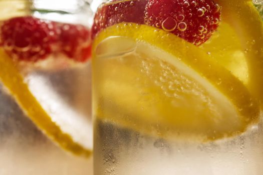 close-up of two glass jars with cold water, lemon slices and red berries, citrus summer refreshments background, copy space