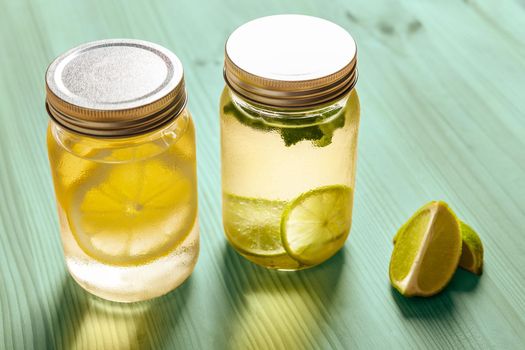 two glass jars with lids full of water with slices of lime and lemon, are on a turquoise wooden table and illuminated by the sunlight, there are also two slices of lime on the table