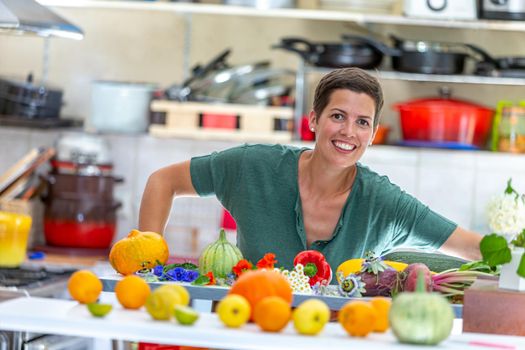 Smiling and relaxed woman, with a bunch of organic vegetables in front of her with the kitchen utensils in blur at the back.