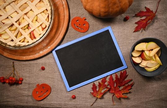 Flat lay composition. Still life with a black board with copy space for advertising text, maple autumn leaves and partial view of a fresh pumpkin pie for Thanksgiving dinner, on a linen tablecloth