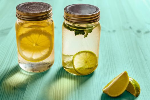 cold summer refreshments with slices of citrus and mint leaves, are in glass jars with lids and illuminated by sunlight, on a turquoise wooden table, there are also lemon wedges