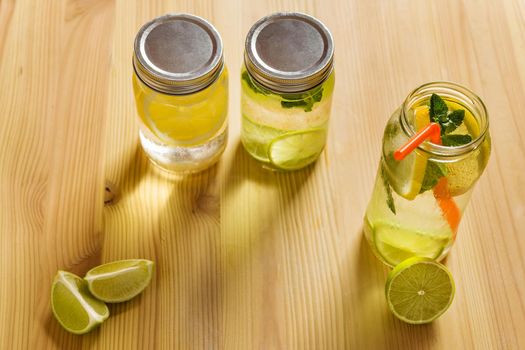 top view of three refreshing lemonades for summer in glass jars, are on a wooden table and illuminated by sunlight, there are pieces of lemon, lime and mint leaves. Image with copy space