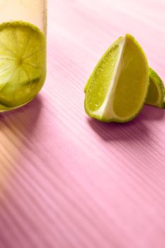 vertical photo with copy space of some slices of lime next to a glass jar with lemonade, this one is illuminated by sunlight and on a pink wooden table