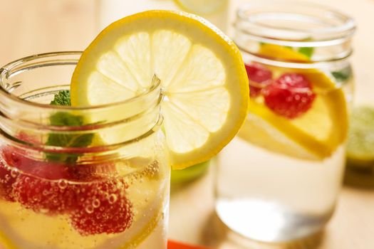 Two glass jars and a bottle with refreshing cold water, slices of lemon, red berries and mint on a wooden table with pieces of citrus lit by sunlight. Summer refreshment background. copy space