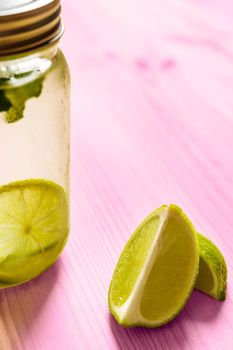 vertical photo of a glass jar with a lid full of lemonade next to some slices of lime, the glass is illuminated by sunlight and on a pink wooden table