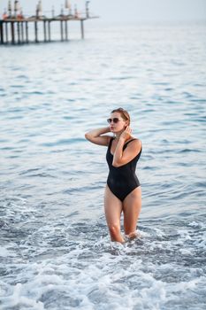 Portrait of a beautiful woman in a black swimsuit with blond hair posing on the beach by the sea. Young woman walking on the beach