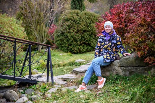 Carefree young woman in trendy vintage pants sitting on table in park and laughing. Curly cute girl in good mood posing in autumn day, enjoying good weather.