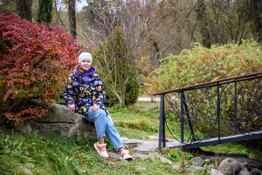 Carefree young woman in trendy vintage pants sitting on table in park and laughing. Curly cute girl in good mood posing in autumn day, enjoying good weather.