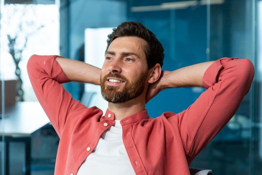Young successful businessman startuper rests in office, man in red shirt throws hands behind head and dreams visualizes, designer thinks about new project.