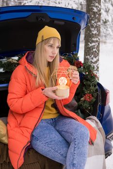 A woman in a winter snow-covered forest in the trunk of a blue car decorated with Christmas decor. The concept of Christmas and winter holidays.