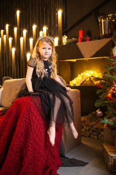 A girl in a festive outfit on the sofa with her dog next to a Christmas tree decorated with garlands, balloons and Christmas toys. The concept of winter holidays is Christmas and New Year holidays. Magical festive atmosphere.