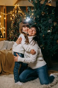 A little girl with her mother in a cozy home environment on the sofa next to the Christmas tree. The theme of New Year holidays and festive interior with garlands and light bulbs.