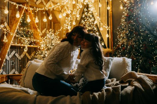 A little girl with her mother in a cozy home environment on the sofa next to the Christmas tree. The theme of New Year holidays and festive interior with garlands and light bulbs