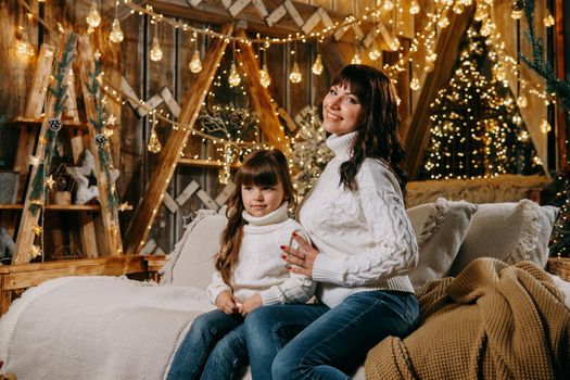 A little girl with her mother in a cozy home environment on the sofa next to the Christmas tree. The theme of New Year holidays and festive interior with garlands and light bulbs.