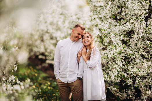 A happy young couple in love stands in a garden of blooming apple trees. A man in a white shirt and a girl in a white light dress are walking in a flowering park