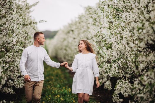 Happy family couple in spring blooming apple orchard. Young couple in love enjoy each other while walking in the garden. The man holds the woman's hand. Family relationships