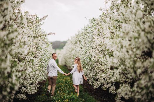 Happy family couple in spring blooming apple orchard. Young couple in love enjoy each other while walking in the garden. The man holds the woman's hand. Family relationships