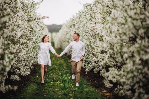 Happy family couple in spring blooming apple orchard. Young couple in love enjoy each other while walking in the garden. The man holds the woman's hand. Family relationships