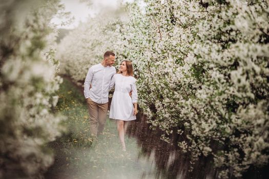 Happy family couple in spring blooming apple orchard. Young couple in love enjoy each other while walking in the garden. The man holds the woman's hand. Family relationships