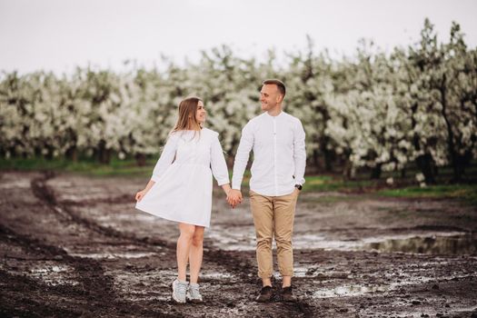 Happy family couple in the spring blooming apple orchard. Young couple in love enjoy each other while walking in the garden. Mud underfoot. The man holds the woman's hand. Family relationships