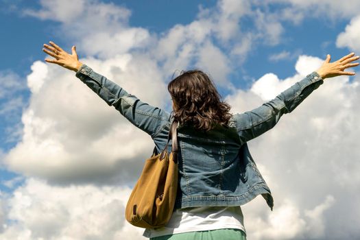 A middle-aged woman in a denim jacket with dark hair happily spread her hands against the sky in sunny weather. positive mood, sense of freedom, joy of life
