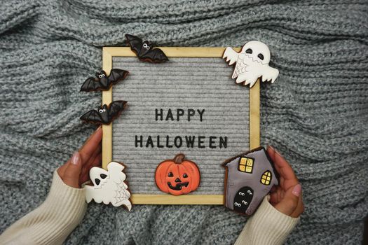 Women's hands hold a board with the text - Happy Halloween. Knitted background. Gingerbread cookies for Halloween celebration.