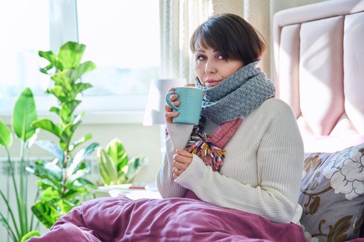 Middle aged woman sitting in bed under blanket warming herself with scarf and hot drink in mug, home lifestyle in cold autumn winter season