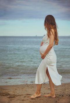 A pregnant woman in a dress looks at the sea. Vertical photo.