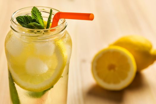 Close-up of the top of a glass jar with lemonade on a wooden table and some unfocused lemons in the background. The jug contains water, slices of lemon, ice, mint leaves and a cane to drink