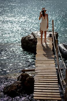 Brunette woman with hat and light beige dress walking on walkway over the sea