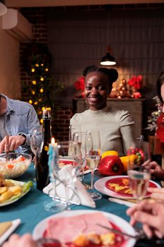 African american woman sitting at christmas festive table portrait, celebrating winter holiday with friends, smiling, looking at camera. Happy girl eating traditional dishes at xmas home party