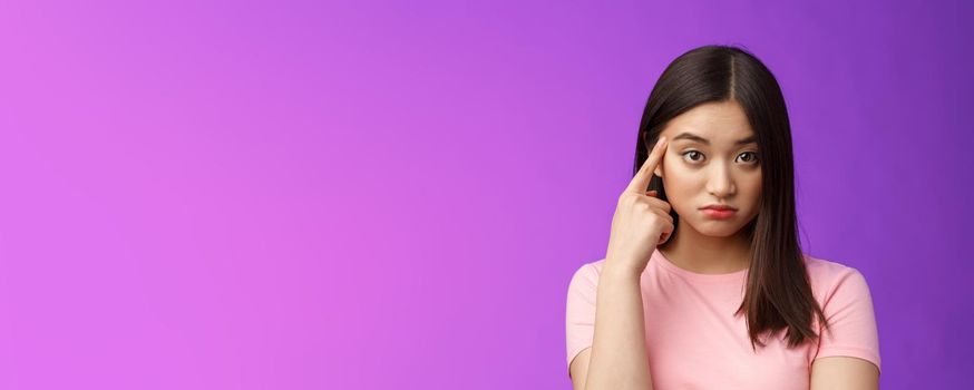 Shocked asian girl scold friend unacceptable behaviour, pointing finger temple look confused and upset stare camera displeased weird actions, asking are you stupid, stand purple background.