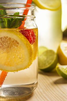 glass jar and bottle lit by sunlight with refreshing cold lemonade water, lemon slices, red berries, mint leaves and drinking canes on a wooden table. Summer citrus soda background, vertical photo