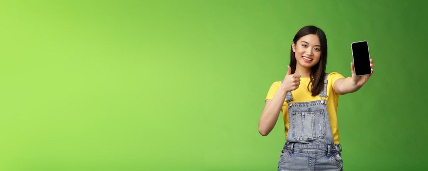 Cheerful cute asian woman judging good smartphone game, show thumb-up like sign, extend arm hold telephone screen, present awesome app, promoting application, stand green background.
