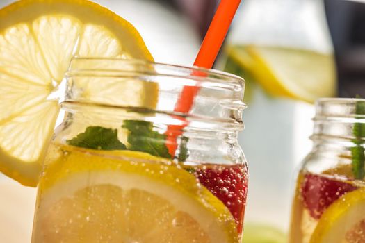 close-up of a glass jar lit by sunlight with refreshing cold lemonade water, lemon slices, red berries, mint leaves and drinking cane. Summer citrus soda background