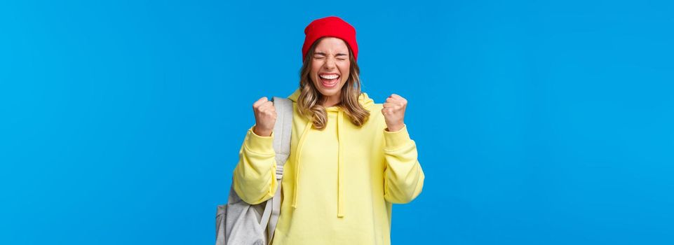Happy cheerful blond girl in red beanie and yellow hoodie, close eyes relieved and smiling triumphing, celebrating great news, passed exams in university, fist pump like champion, winning prize.