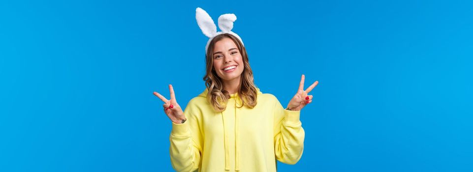 Holidays, traditions and celebration concept. Kawaii young blond girl in rabbit ears showing peace gesture and smiling, having fun, enjoying party, standing blue background.