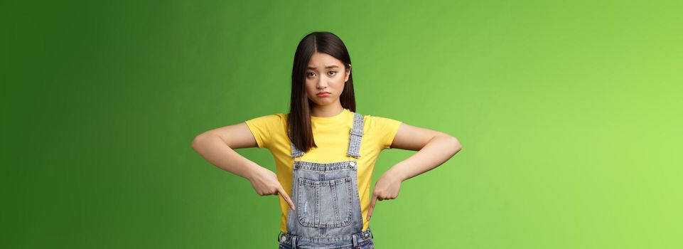 Sad asian girl in dispair feel upset, frowning sulking pessimistic, pointing down look camera pulling sorrow uneasy face, express sadness and frustration, stand green background. Copy space