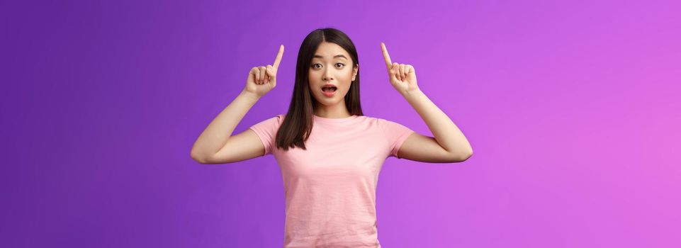 Excited cute surprised asian girl in pink t-shirt telling about awesome news pointing raised fingers up, indicate top advertisement, open mouth astonished, thrilled stand blue background.